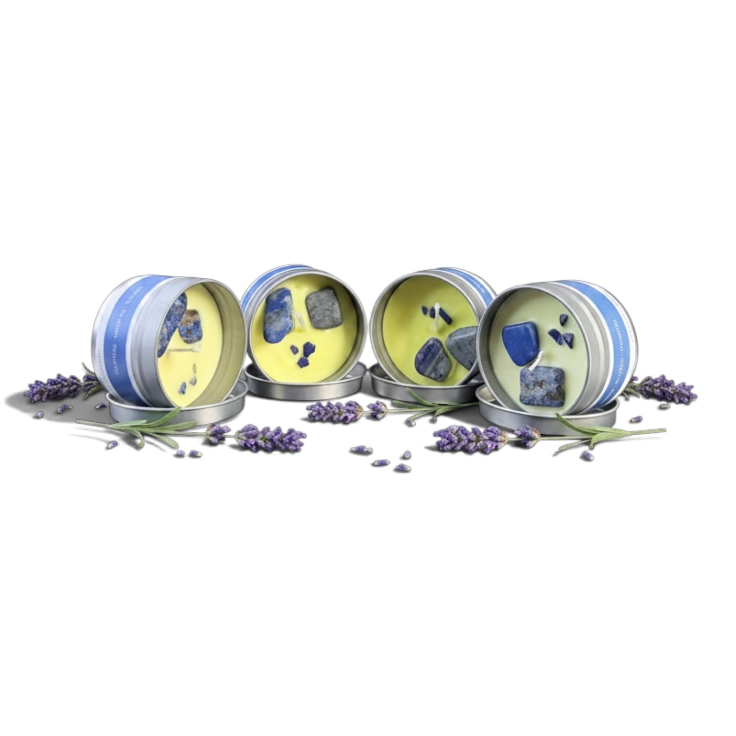 Four round metal tins of lavender soap, arranged side by side on a white background with scattered lavender sprigs. The tins have blue and white striped borders, silver lids, and pale yellow soap inside, each decorated with blue and gray stone inclusions.